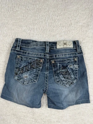 Blå jeansshorts från Miss Me - Säljer ett par blå jeansshorts från Miss Me med snygga kontrastsömmar och dekorativa knappar. Klassisk femficksmodell med bälteshällor och dragkedja. Perfekta för sommaren och ger en avslappnad men trendig look.