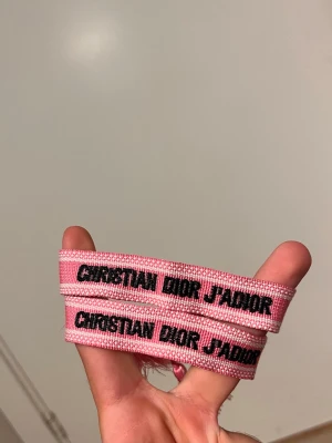 Christian Dior rosa tygarmband - Två rosa tygarmband från Christian Dior med texten 'J'ADIOR' i svart. Armbanden har en vävd struktur och är dekorerade med små vita detaljer längs kanterna. Perfekt accessoar för att lyfta din outfit.