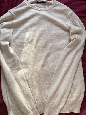 Beige stickad tröja från J.Lindeberg - Snygg beige stickad tröja från J.Lindeberg i tunn ullmix. Tröjan har rund halsringning, långa ärmar och diskret logga på bröstet. Perfekt för lager på lager och enkel att matcha med olika outfits.