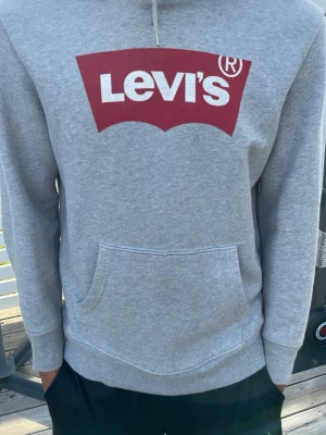 Grå Levi's hoodie med röd logga - Snygg grå hoodie från Levi's med stor röd logga på bröstet. Klassisk känguruficka framtill och huva med dragsko. Tillverkad i mjukt sweatshirtmaterial som är perfekt för chill dagar. Långärmad och enkel att matcha med det mesta.
