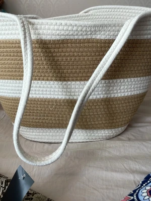 Randig shoppingväska i beige och vitt - Snygg shoppingväska med breda ränder i beige och vitt. Väskan har två långa handtag och är tillverkad i ett flätat, textilliknande material. Perfekt storlek för att få plats med allt du behöver. Enkel och stilren design som passar till många outfits.