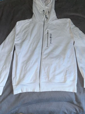 Vit hoodie från Sail Racing - Säljer en vit hoodie från Sail Racing med dragkedja framtill och en bröstficka med zip. Hoodien har logga broderad på bröstet och justerbar huva med snören. Tillverkad i mjukt bomullsmaterial och har ribbade muddar vid ärmslut och nederkant.