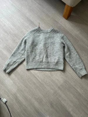 Ljusgrå stickad tröja - En stilren ljusgrå stickad tröja med rund hals och långa ärmar. Tröjan har ribbstickade muddar vid ärmslut och nederkant samt en något boxig passform. Perfekt för lager på lager och enkel att matcha med olika outfits.