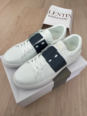 Valentino Garavani vita sneakers - Snygga vita sneakers från Valentino Garavani med bred svart rem över mitten och vita skosnören. Tillverkade i slätt läder med guldfärgad logga på plösen. Klassisk låg modell som passar till många olika stilar.