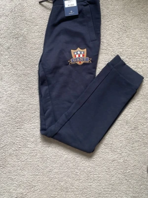 Mörkblå mjukisbyxor från GANT - Mörkblå mjukisbyxor från GANT med broderad logga på benet. Byxorna har ribbade muddar vid benslut och resår i midjan med dragsko. Tillverkade i mjukt material för maximal komfort och har praktiska sidofickor. Helt oanvända.