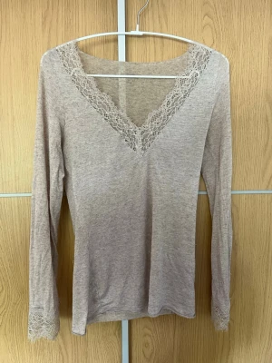Beige långärmad från intimismi  med spets - Säljer en beige långärmad topp med v-ringning och fina spetsdetaljer vid halsringningen och ärmsluten. Toppen har en slim passform och är tillverkad i ett mjukt material som känns skönt mot huden. Perfekt för dig som gillar en enkel men söt stil. Bra att veta, några trådar som hänger samt ett litet hål i den