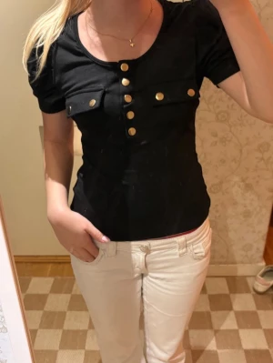 Svart topp med guldknappar - Snygg svart topp med korta ärmar och dekorativa guldknappar framtill. Toppen har två fickor på bröstet och en figurnära passform som framhäver siluetten. Perfekt att styla med både jeans och kjol🤩❣️