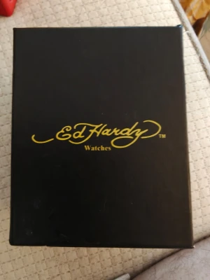 Ed Hardy ~Love Kills Slowly~ - Säljer en Ed Hardy klocka med stort dödskallemotiv och texten 'Love Kills Slowly' på urtavlan. Klockan har en bred svart rem i skinn och silverfärgat boett. Urtavlan pryds också utav Swarovski kristaller. Perfekt för dig som gillar statement-accessoarer och unik design. Remmen lever nog ett tag till fick smörja upp den med fett så den fick lite mobilitet tillbaka. 