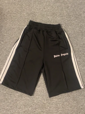 Svarta Palm Angels shorts med vita ränder - Säljer ett par svarta shorts från Palm Angels med vita sidoränder och logga på vänster ben. Shortsen har elastisk midja och sportig look, perfekta för en avslappnad stil. Materialet känns mjukt och lätt, troligen polyester.