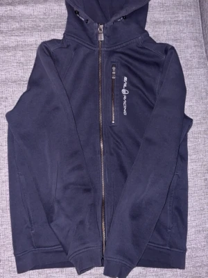 Mörkblå hoodie från Sail Racing - Mörkblå hoodie från Sail Racing med dragkedja framtill och en bröstficka med logga. Hoodien har huva med dragsko och är tillverkad i mjukt material, perfekt för en avslappnad stil. Snygga detaljer och klassisk passform.