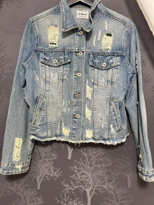 Ljusblå distressed jeansjacka G. Cause - Trendig ljusblå jeansjacka från G. Cause med slitna detaljer och rå kant nertill. Klassisk knappstängning, två bröstfickor med lock och två sidofickor. Perfekt för dig som gillar en avslappnad och edgy stil.
