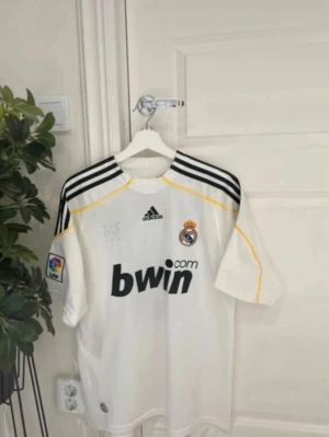 Real Madrid Ronaldo matchtröja - Vit Real Madrid matchtröja från Adidas med svarta detaljer och gula inslag. Tryck med 'Ronaldo' och nummer 9 på ryggen. Bwin-logga på bröstet och La Liga-märke på ärmen. Perfekt för fotbollsfans som vill sticka ut på planen eller läktaren.