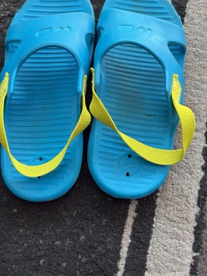 Blå sandaler med gula remmar - Säljer ett par blå sandaler med gula textilremmar. Sandalerna är gjorda i lätt plastmaterial och har räfflad sula för extra grepp. Perfekta för stranden eller poolen och enkla att ta på och av.