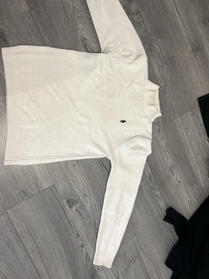 Vit stickad tröja med logga - Klassisk vit stickad tröja med rund hals och lång ärm. Tröjan har ribbade muddar vid ärmslut och nederkant samt en liten mörk broderad logga på bröstet. Perfekt för lager på lager och enkel att matcha med olika stilar.