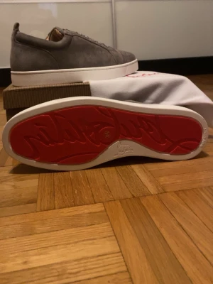 Gråa sneakers med röd sula - Stilrena gråa sneakers i mocka från Christian Louboutin med den ikoniska röda sulan. Skorna har vita, tjocka sulor och klassisk snörning framtill. Perfekta för dig som vill ha en clean look med en twist.