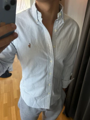 Randig skjorta från Ralph Lauren - Klassisk vit skjorta med tunna blå ränder från Ralph Lauren. Skjortan har lång ärm, knappar framtill och button-down krage. Broderad logga på bröstet. Perfekt för en clean och stilren look.