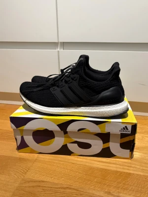 Svarta Adidas Ultraboost sneakers - Snygga svarta Adidas Ultraboost sneakers med stickad ovandel och klassiska tre ränder på sidan. Skon har en vit Boost-mellansula för extra komfort och svart yttersula med grepp. Perfekt för dig som gillar stilrena och bekväma sneakers. Lite trasig låda