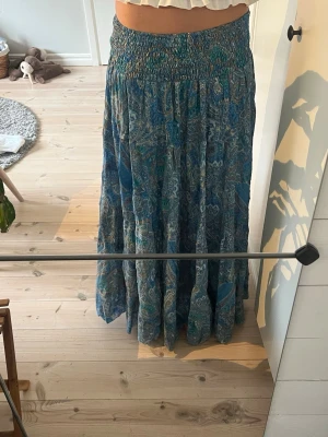 Blå långkjol med paisleymönster - Säljer en flowy blå långkjol med paisleyinspirerat mönster i olika blå och gröna toner. Kjolen har smock i midjan för skön passform och är gjord i ett tunt, luftigt material som passar perfekt till varmare dagar. Köpt på en marknad utomlands💞