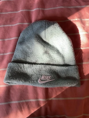 Grå ribbstickad mössa från Nike - Snygg grå ribbstickad mössa från Nike med uppvikt kant och broderad Nike-logga i rosa framtill. Klassisk beanie-modell som passar perfekt till höst och vinter. Tillverkad i mjukt material för skön känsla.