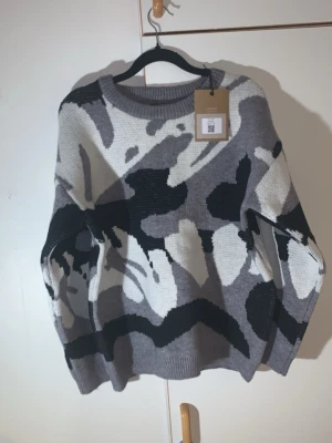 S. Camo grå stickad  - Köpte den va för lite på lillebror tänkte ville ha lite tight fit han har m i verkligheten modellen är redan bra fit Säljer en stickad tröja i olika nyanser av grått, svart och vitt med ett coolt abstrakt mönster. Tröjan har rund halsringning, långa ärmar och ribbade muddar vid ärmslut och nederkant. Perfekt för dig som gillar unik design och sköna material.