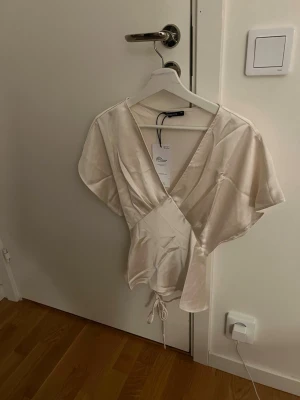 Champagnefärgad omlottopp i satin - Snygg champagnefärgad omlottopp i satin med korta ärmar och knyt i midjan. Toppen har en djup v-ringning och ett elegant, glansigt material som ger en lyxig känsla. Perfekt att styla med jeans eller kjol för en trendig look.