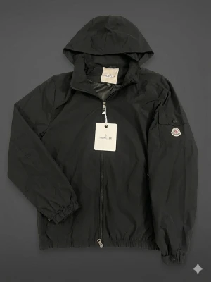 Svart vindjacka från Moncler - Snygg svart vindjacka från Moncler med huva och dragkedja framtill. Jackan har elastiska muddar vid ärmsluten och en ficka med lock och Moncler-logga på ärmen. Tillverkad i lätt och slitstarkt material, perfekt för blåsiga dagar.