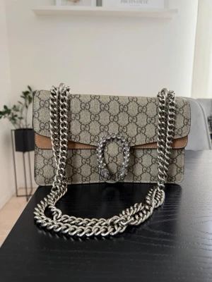 Axelremsväska med kedja - Snygg axelväska i beige canvas med klassiskt GG-mönster och detaljer i brunt läder. Väskan har en robust silverfärgad kedja och ett dekorativt spänne framtill. Insidan är fodrad med GG-tyg och har magnetlås. Perfekt för att lyfta din outfit.