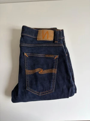 Mörkblå jeans från Nudie Jeans - Snygga mörkblå jeans från Nudie Jeans med klassisk femficksmodell och kontrastsömmar i orange. Jeansen har en rak passform och är tillverkade i denim av bomull. Perfekta för dig som gillar stilrena och tidlösa jeans. Storlek 31/32 modell, lean Dean
