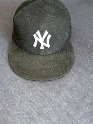 Svart New Era Yankees keps - Snygg svart keps från New Era med klassisk New York Yankees-logga broderad i vitt framtill. Kepsen har rak skärm och ventilationshål upptill för extra komfort. Materialet är tåligt tyg som passar perfekt till streetwear-stilen.