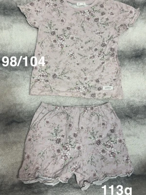 Blommig pyjamas i ljusrosa - Söt tvådelad pyjamas med kortärmad topp och shorts i ljusrosa med blommigt mönster. perfekt för mysiga kvällar. Volangdetaljer vid ärmslut och benslut ger en extra gullig touch.🌸Två svaga fläckar. Storlek 98/104