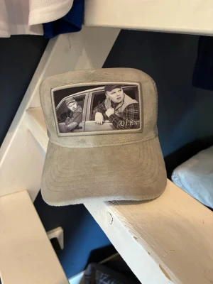 Grå velvet keps Leif och Billy - Säljer en grå velvet keps med mesh baktill och justerbar snapback. Framtill finns ett svartvitt fototryck av två personer i en bil, vilket ger kepsen en unik och retro vibe. Perfekt för dig som vill sticka ut med din stil. Eget företag som lagt ned