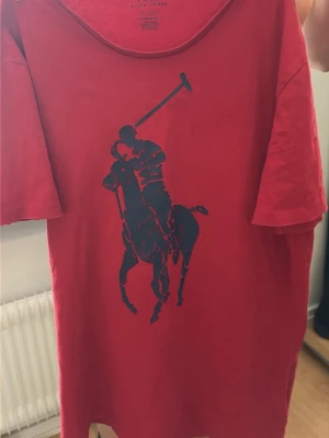 Röd t-shirt Ralph Lauren  - Snygg röd t-shirt från Ralph Lauren med stort blått Polo-tryck på framsidan. Klassisk rund halsringning och korta ärmar. Perfekt för en avslappnad och sportig stil.