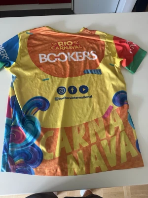 Färgglad Rio Carnaval t-shirt - Unik t-shirt med Rio Carnaval-tema och färgstarkt tryck över hela plagget. Framsidan har ett stort motiv av en kvinna med glasögon och huvudscarf, samt texten 'RIO CARNAVAL'. T-shirten har rund hals och korta ärmar i olika färger och materialet känns lätt och syntetiskt.