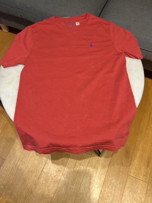 Röd t-shirt med blå logga - Säljer en röd t-shirt med korta ärmar och en liten blå logga broderad på bröstet. T-shirten har en klassisk passform och rund halsringning. Perfekt till jeans eller shorts för en enkel och snygg look.