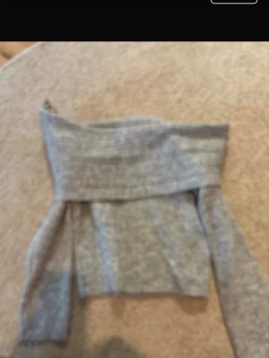 Grå off shoulder stickad tröja - Trendig grå stickad tröja med off shoulder-design och bred ribbad kant upptill. Tröjan har långa ärmar och är gjord i mjukt, ribbstickat material. Perfekt för dig som gillar en avslappnad men ändå stilren look.