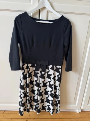 Svart och vit klänning Rinascimento - Snygg svart och vit klänning från Rinascimento med trekvartsärm och rund hals. Överdelen är svart och kjolen har ett stort, rutigt mönster i svart och vitt samt en liten rosett i midjan. 