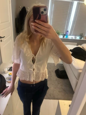 Vit blus med snörning och spetsdetaljer - Söt vit blus med korta ärmar, snörning framtill och broderade spetsdetaljer längs kanterna. Modellen är croppad och har volang nedtill för en romantisk vibe. Perfekt till jeans eller kjol för en trendig look.