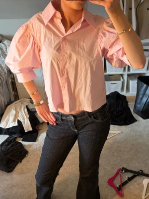 Ljusrosa puffärmad blus - Trendig ljusrosa blus med puffiga korta ärmar och klassisk krage. Blusen har knäppning framtill och är tillverkad i ett lätt, vävt material som ger en luftig känsla. Perfekt för dig som vill ha en söt och stilren look.