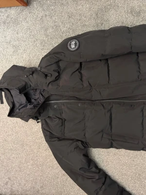 Svart dunjacka från Canada Goose - Säljer en svart dunjacka från Canada Goose med avtagbar huva och dragkedja framtill. Jackan har ett klassiskt patchmärke på ärmen och stora, quiltade paneler för extra värme. Perfekt för kalla vinterdagar.