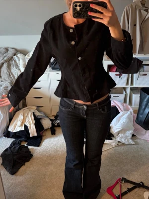 Svart figursydd jacka med knappar - Säljer en svart figursydd jacka med rund halsringning och stora silvriga knappar framtill. Jackan har lång ärm och lätt utsvängd midja för en snygg siluett. Perfekt till jeans eller kjol för en clean look.