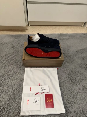 Marinblå Christian Louboutin sneakers - Snygga marinblå sneakers från Christian Louboutin med klassisk röd sula och svarta skosnören. Skorna är låga och har en ovandel i mocka som ger en exklusiv känsla. Perfekta för dig som vill sticka ut med en ikonisk detalj på fötterna. Skorna är helt oanvända, baravteatade. Jag säljer dessa skor då dem är för små på mig.