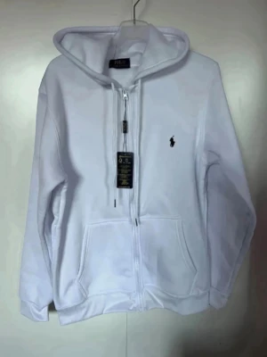 Vit hoodie från Polo Ralph Lauren - Snygg vit hoodie från Polo Ralph Lauren med dragkedja framtill och klassisk logga broderad på bröstet. Hoodien har huva med dragsko och två stora fickor framtill. Perfekt för en clean och stilren look. Betalning genom vanlig swish! 