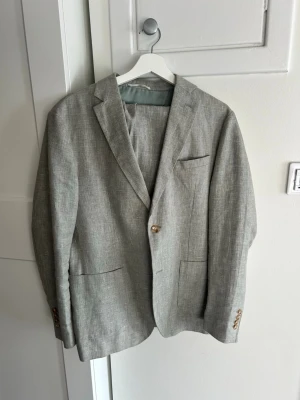 Grön linnekostym John Henric - Stilren grön kostym i linne med två knappar framtill och klassisk passform. Kavajen har tre fickor fram, slits bak och dekorativa knappar vid ärmsluten. Perfekt för en avslappnad men ändå uppklädd look.