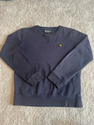 Mörkblå stickad tröja Lyle & Scott - Mörkblå stickad tröja från Lyle & Scott med klassisk rund halsringning och ribbade muddar. Tröjan har det ikoniska gula fågelmärket broderat på bröstet. Perfekt för dig som gillar stilrena och tidlösa plagg.
