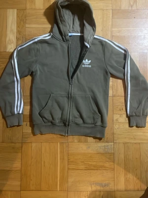 Olivgrön Adidas hoodie med dragkedja - Säljer en olivgrön hoodie från Adidas med dragkedja framtill och vita ränder längs ärmarna. Klassisk Adidas-logga på bröstet och två fickor fram. Hoodien har ribbade muddar och huva. Perfekt för en avslappnad streetwear-look.
