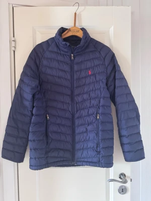 Mörkblå pufferjacka från Polo Ralph Lauren - Snygg mörkblå pufferjacka från Polo Ralph Lauren med diskret röd logga på bröstet. Jackan har dragkedja framtill, två sidofickor med dragkedja och en hög krage. Stl S (14-16) ena fickans dragkedja sönder 
