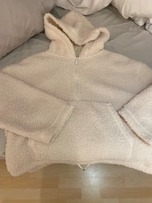 Vit teddyjacka med huva och dragkedja - Supermjuk och fluffig teddyjacka i offwhite med stor huva och halv dragkedja framtill. Jackan har en rymlig känguruficka och avslappnad passform. Perfekt för kalla dagar när du vill vara både varm och stilren.