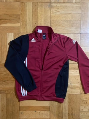 Vinröd och svart Adidas träningsjacka - Snygg vinröd träningsjacka från Adidas med svarta ärmar och sidor. Jackan har dragkedja framtill, vita Adidas-loggor på bröst och ärm samt klassiska vita ränder på sidan. Tillverkad i ett lätt och stretchigt material, perfekt för träning eller uppvärmning.