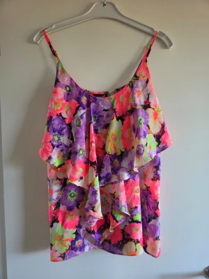 Blommig ärmlös linnetopp volang - Färgstark linnetopp med blommigt mönster i rosa, lila, gul och grön. Toppen har tunna axelband, volangdetaljer framtill och är ärmlös. Materialet känns lätt och luftigt, perfekt för varma dagar.