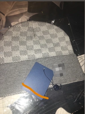 Grå mössa med rutigt mönster - Snygg grå mössa med klassiskt rutigt mönster och bred uppvikt kant. Perfekt accessoar för att hålla värmen och ge en stilren touch till din outfit. Mössan har en diskret logotyp på kanten och är tillverkad i mjukt material.